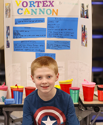  Boy sitting in front of his display at science fair. Display reads "vortex cannon"